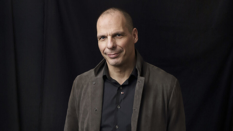 A photo of Yanis Varoufakis standing in front of a black curtain backdrop