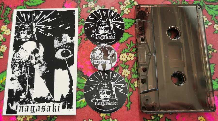 A top down view of the contents of the tape version, from the left: the front cover, two round stickers and a pin with Nagasaki artwork, and the cassette itself, in a brown translucent shell